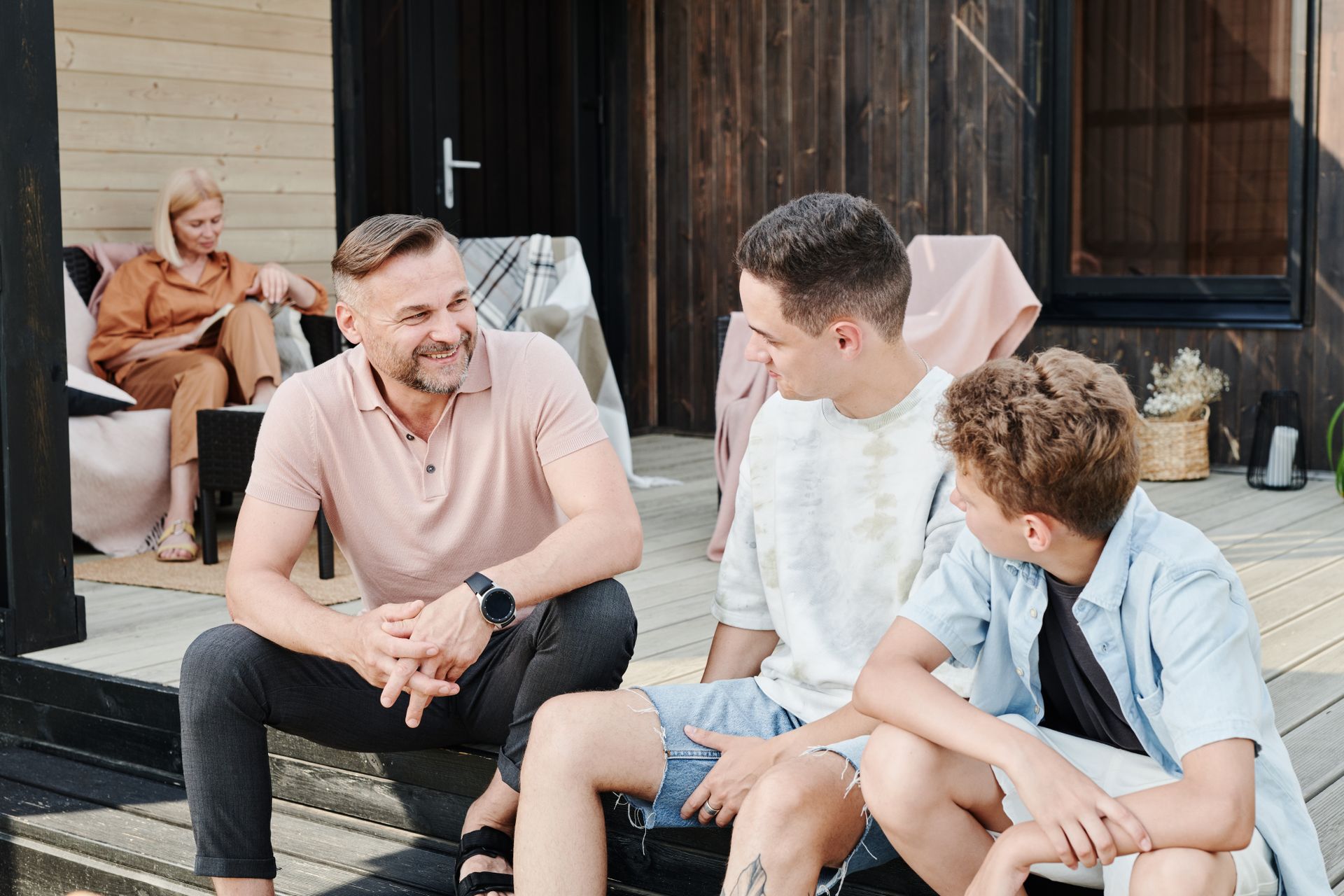A father laughs with his adult son and younger son on a porch step, mother reading quietly behind them.