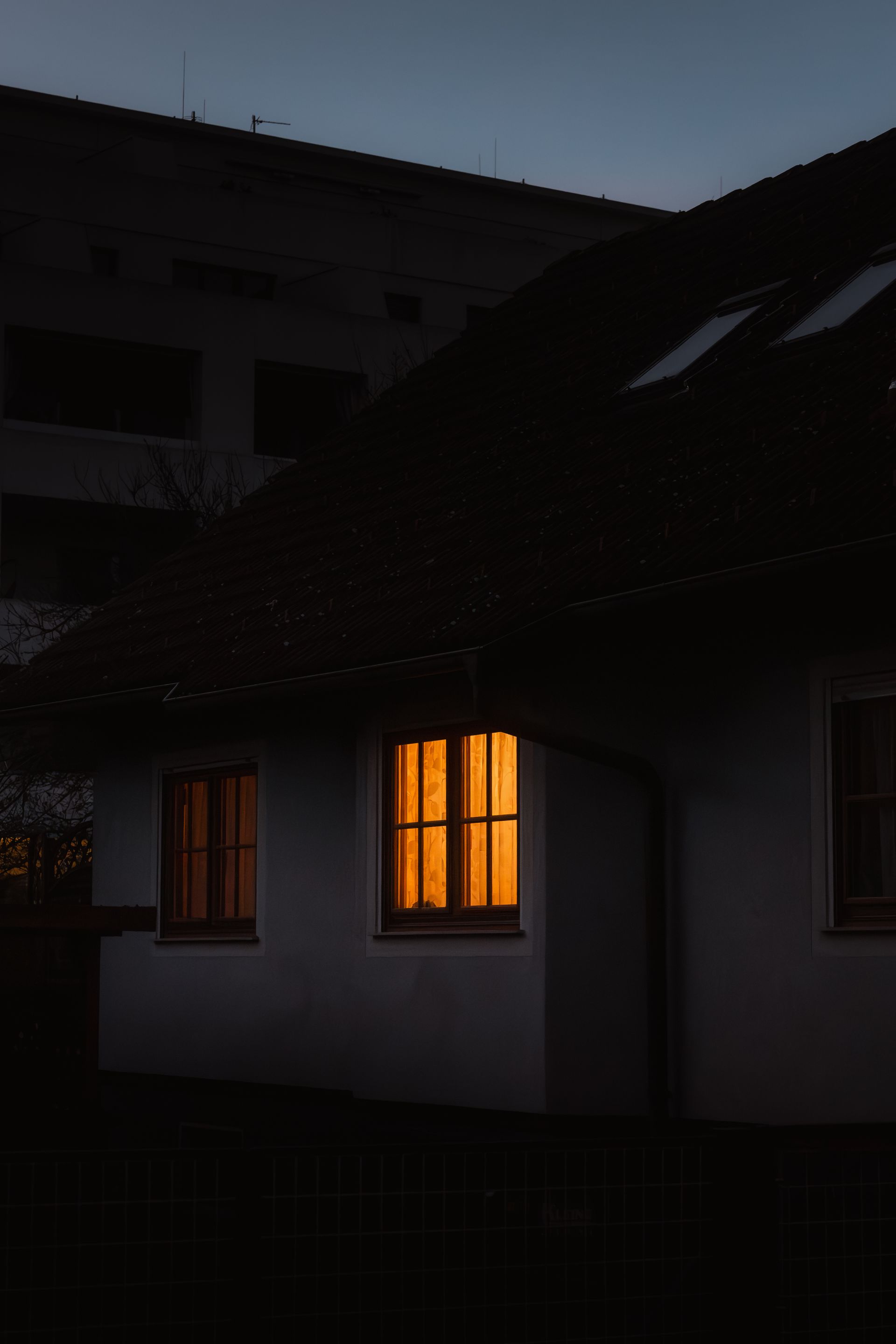 A single lamplit window glowing in an otherwise dark, quiet house at dusk