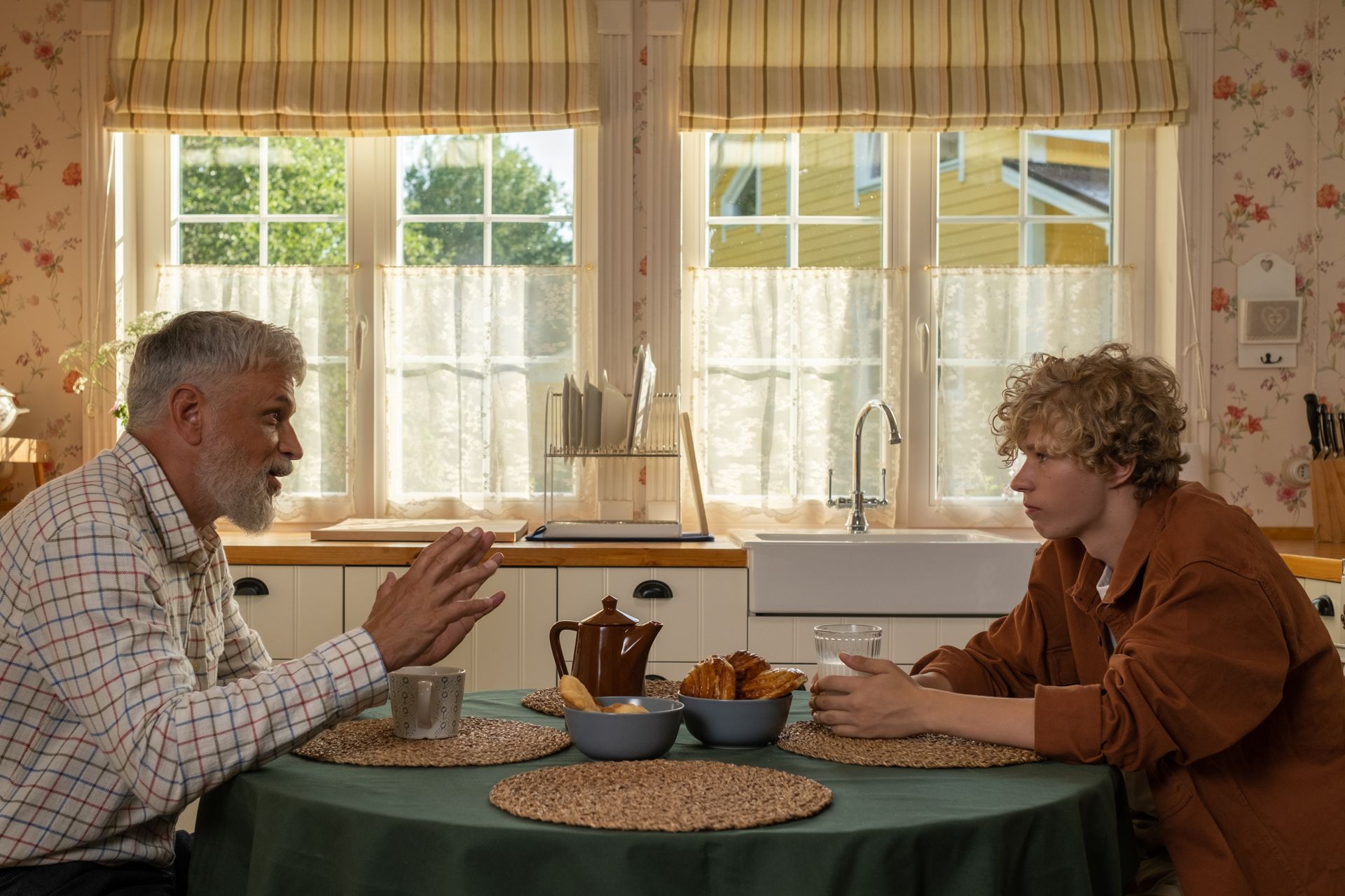 A silver-haired mentor and a young man share an animated, eye-contact conversation over coffee at a sunlit kitchen table.