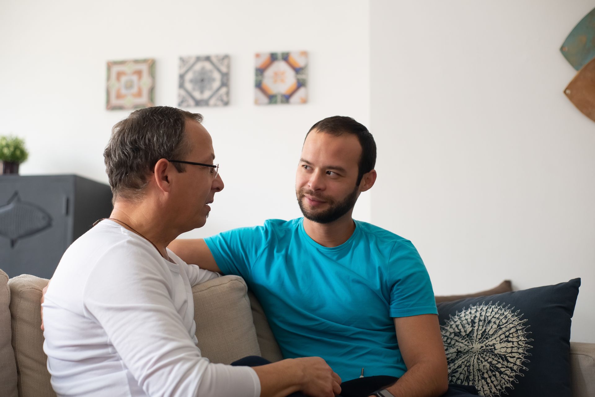 An older mentor and a young man in an unhurried one-on-one conversation on a living-room sofa.