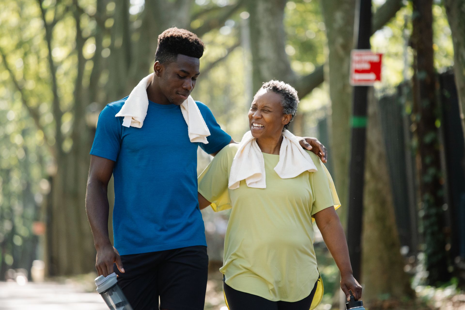 A mother and her adult son walk together along a sunlit, tree-lined path.