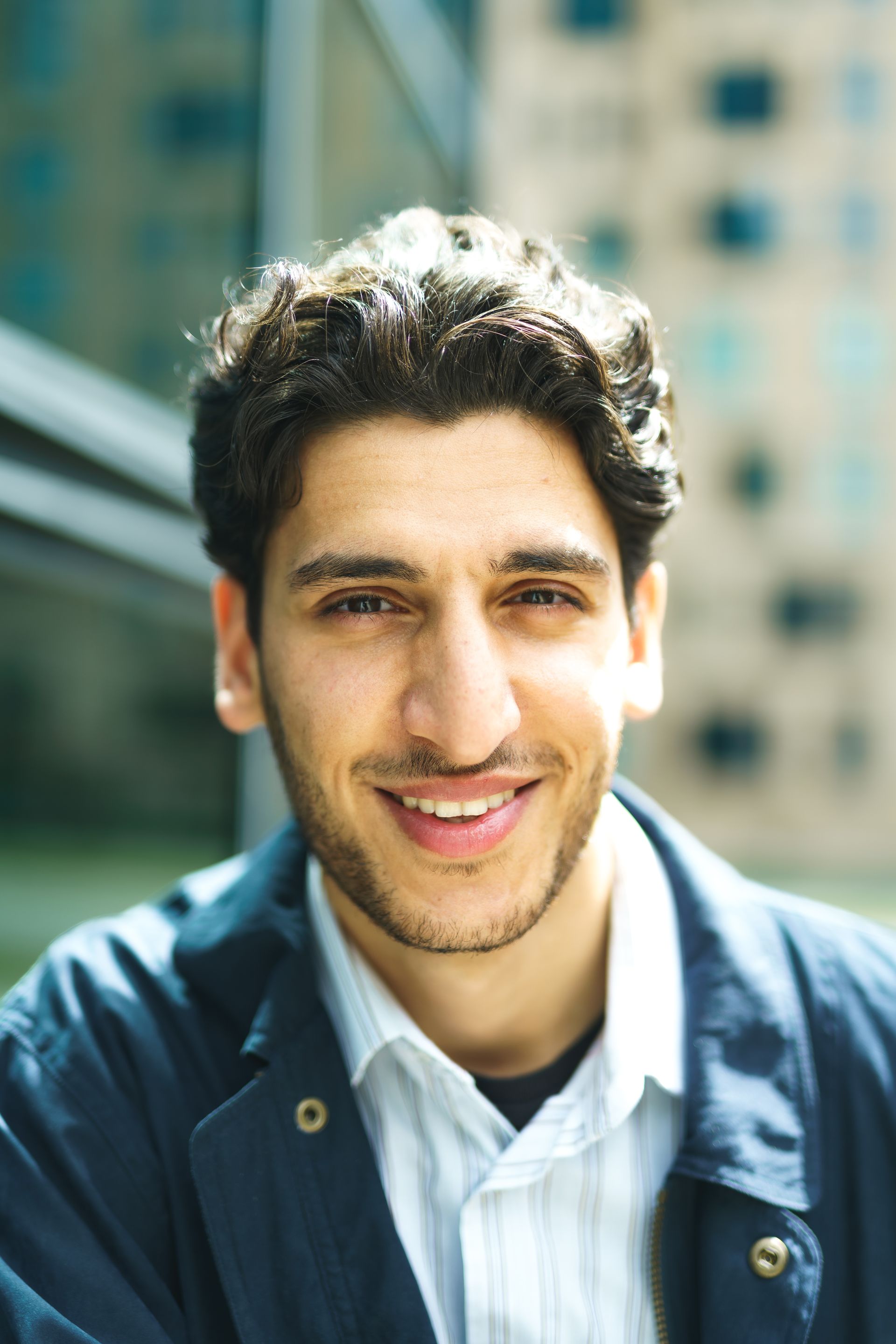 A young man smiling genuinely in bright daylight, soft city bokeh behind him.